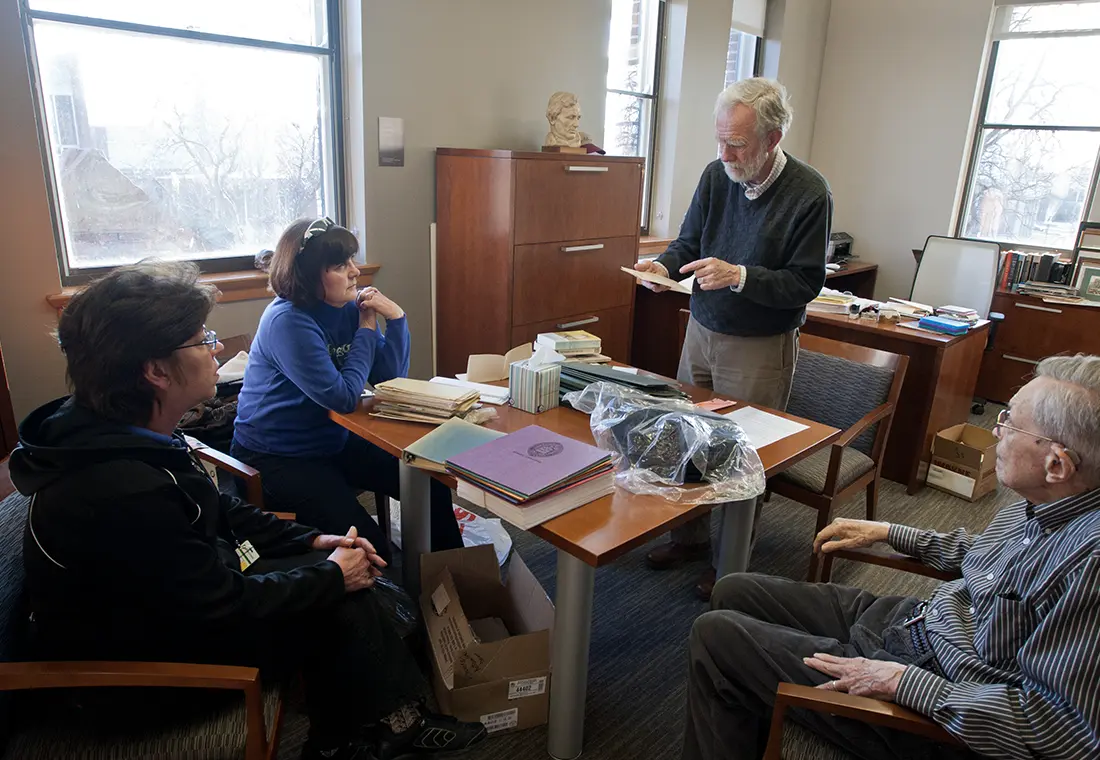 A group of people sit around a table while the person on one side stands and reads from a piece of paper.