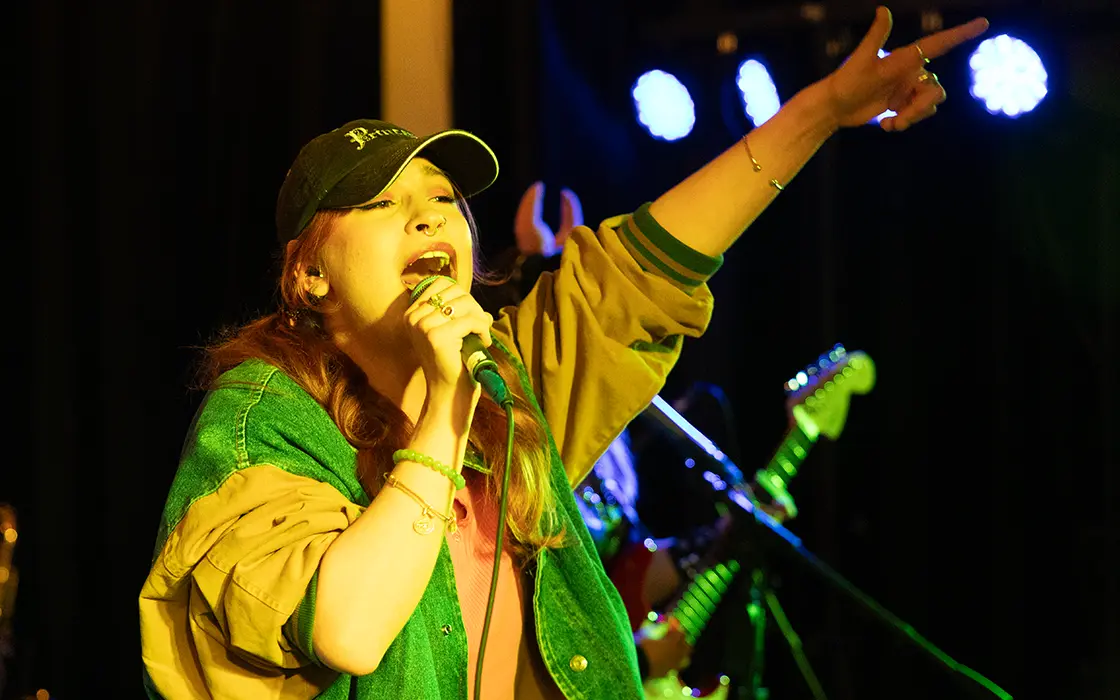 A student in a baseball cap sings and holds her hand into the air.