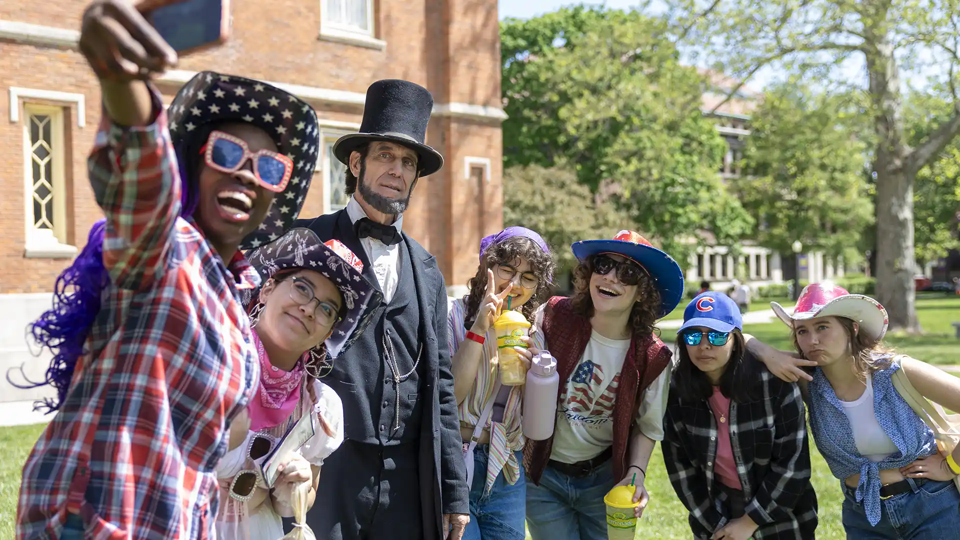 A group of students take a selfie with an Abraham Lincoln impersonator in front of Old Main. Students are wearing sunglasses and cowboy hats.