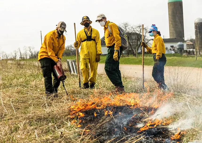 Three students and a professor, all dressed in protective gear, oversee a controlled fire in a field.