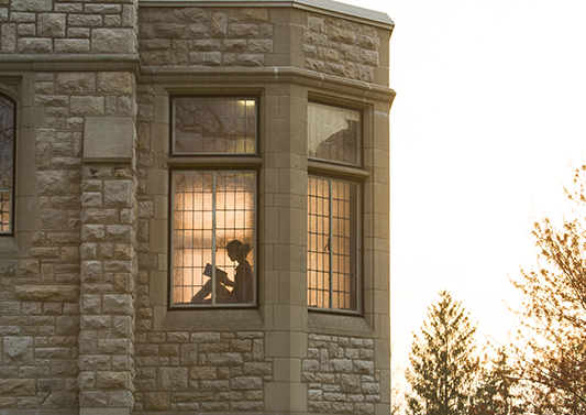 A student is seen reading a book through the library windows. 