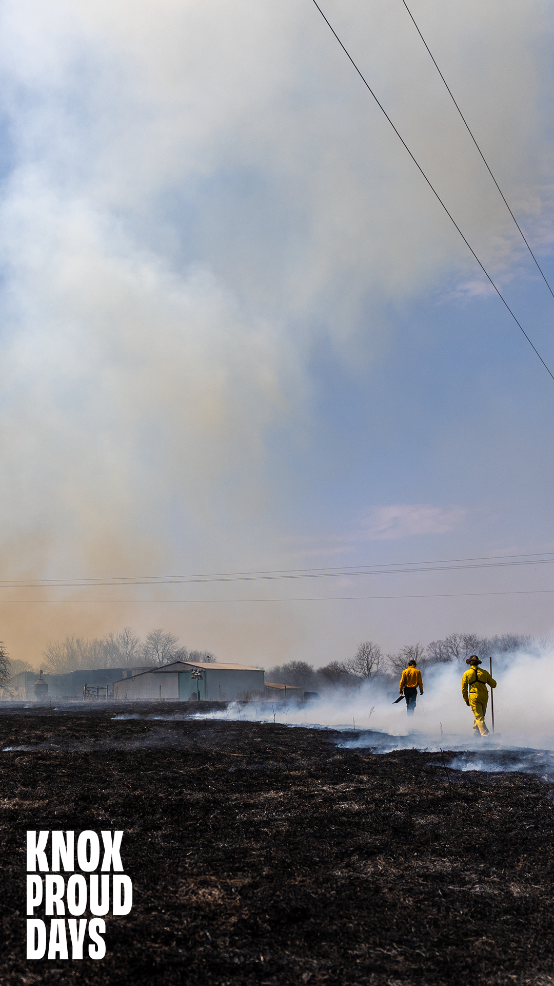 People in yellow gear walk through a field that is smoking. Smoke fills the air above them.