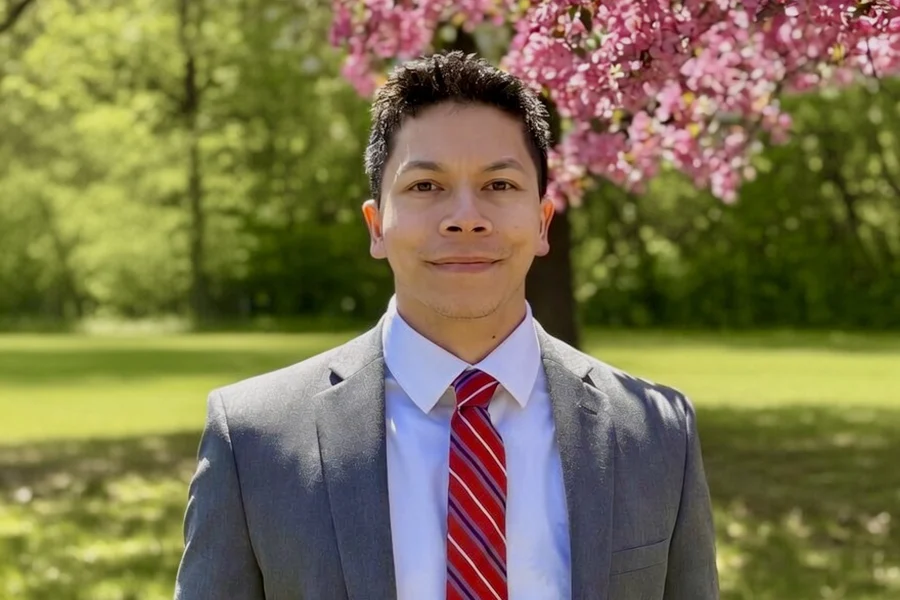 A person wearing a suit and tie smiles at the camera; trees, pink leaves, and grass are visible in the background.