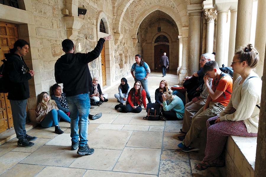 Students and professors sit on the ground and on nearby railings outside an ancient building as their guide points out notable features in its design.