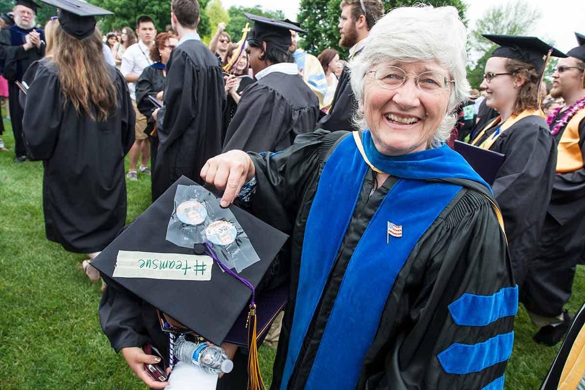 L. Sue Hulett, Richard P. and Sophia D. Henke Distinguished Professor of Political Science, points to a kneeling student's mortarboard art at 2014 Commencement, which reads "#teamsue."