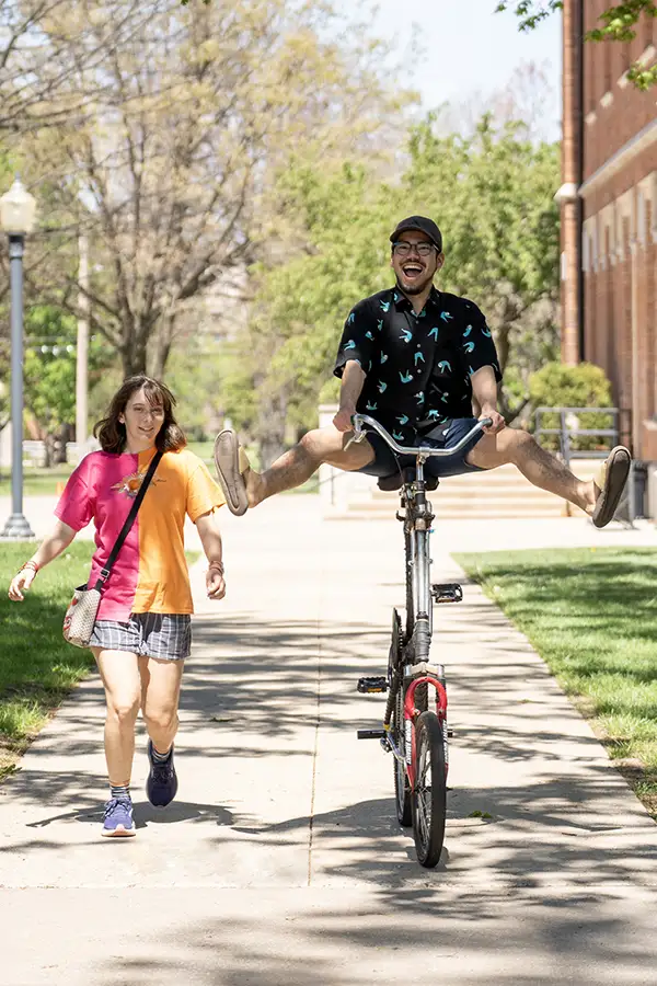 A student rides a tall bike with his legs lifted up in the air. Another student walks alongside him.