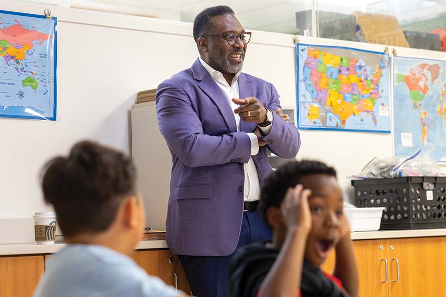 President McGadney stands at the front of a classroom, pointing at a student who has a shocked expression on his face.