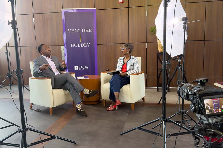 President McGadney and Dorothy Tucker sit in two white chairs facing each other, surrounded by cameras. There is a purple pull-up banner behind them that reads "Venture Boldly."