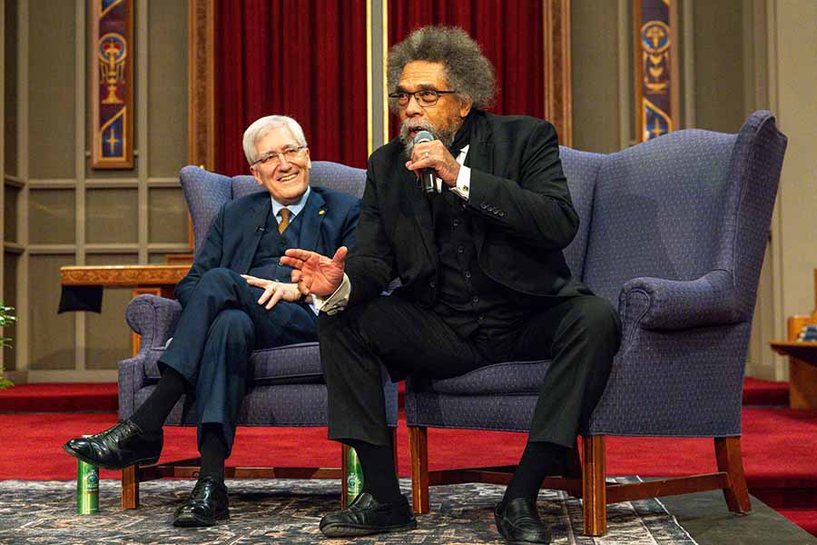 Two men sit next to each other on a stage. One man is speaking into a microphone.