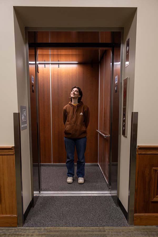 A student stands inside of an elevator, looking up and smiling.