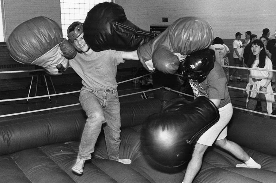 A black and white photo of two students in a boxing ring, boxing each other with large inflatable boxing gloves on.