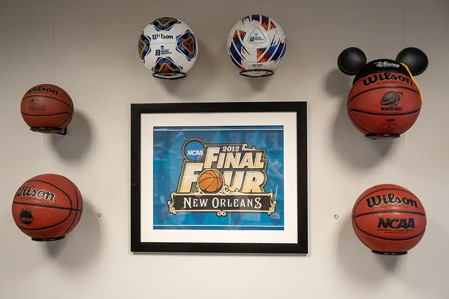 A white wall with six different basketballs and soccer balls hanging there, with the Final Four New Orleans framed poster in the middle of the balls. One basketball has Mickey Mouse ears on it.