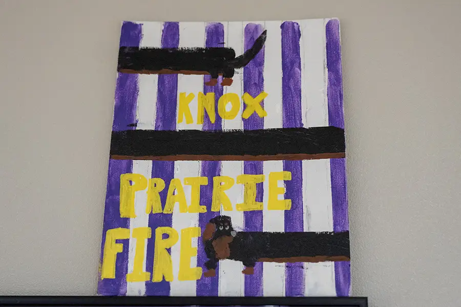 A painting with white and purple vertical lines, and Knox Prairie Fire in yellow with a black dog painted on it. 