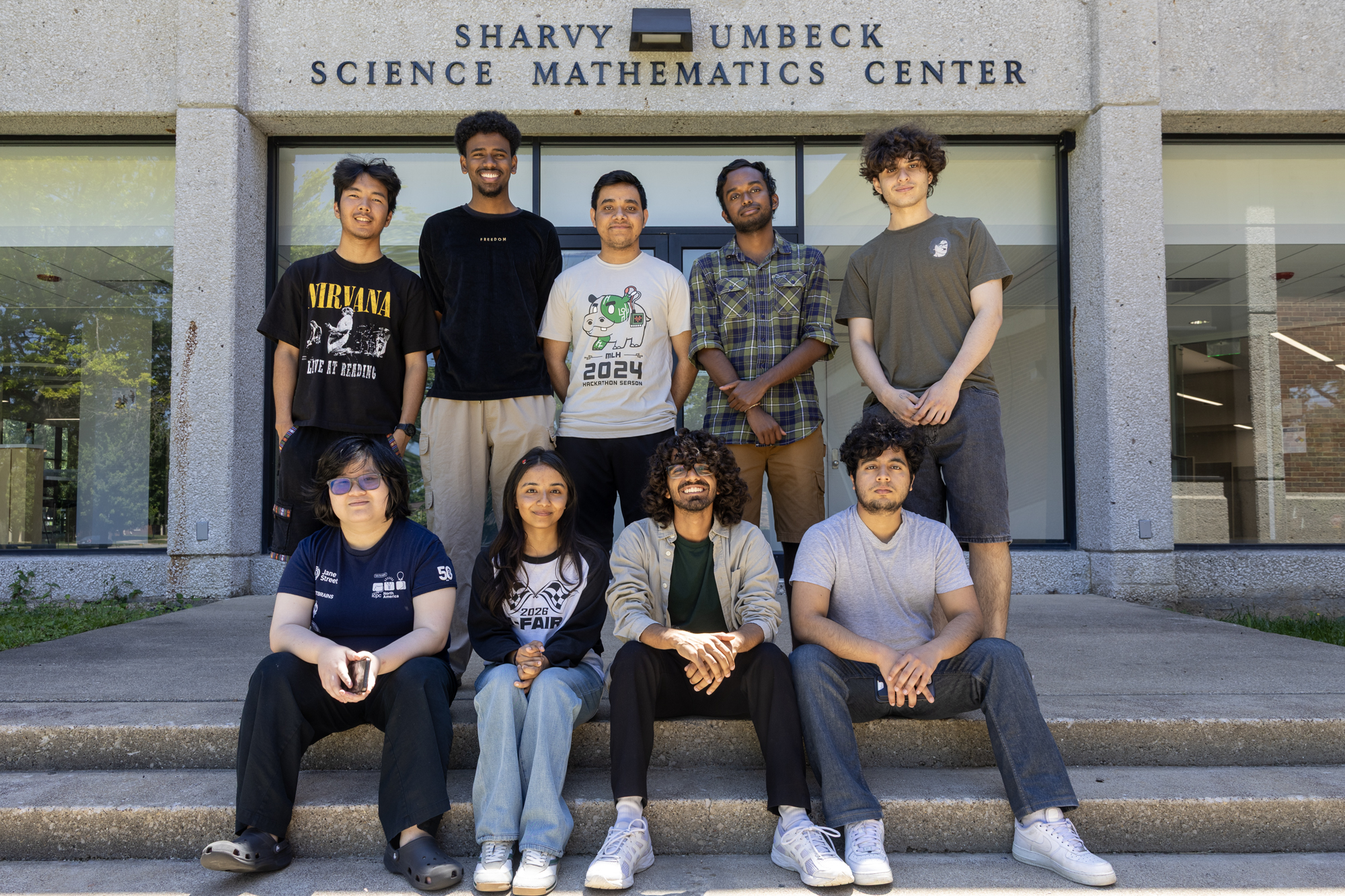 A group of students sit and stand on the steps in front of doors of the Sharvy Umbeck SCience and Mathematics Center.