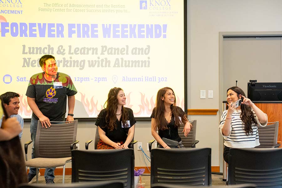 A diverse group of alumni sit at the front of a classroom, smiling and talking.