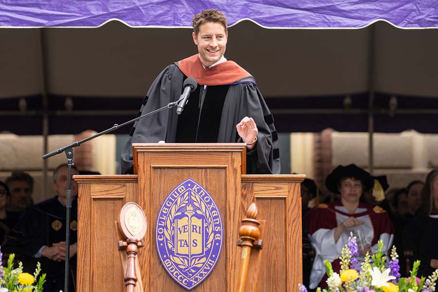 Justin Hartley stands at a podium addressing the graduating class. He is wearing a Commencement gown.
