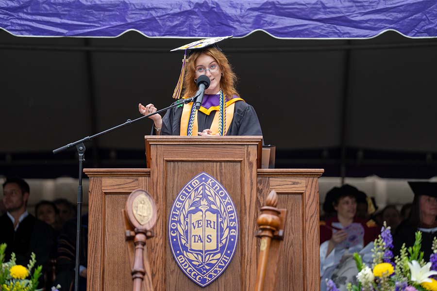 Patience Claire Wagner ’26 stands at a podium outside on a stage, speaking into a microphone while wearing a graduation cap and gown.