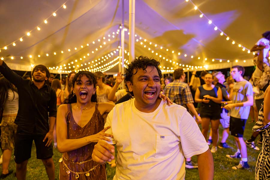 Two people are in the front of a crowd who is dancing underneath an outdoor tent with lights hanging from the ceiling. 