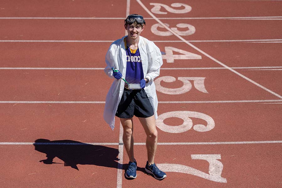 Lance Miller stands on a track, holding laboratory supplies and with laboratory goggles on his head and a lab coat on.