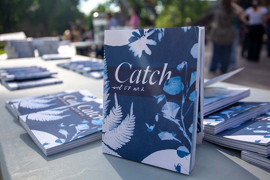 A volume of Catch sits on a table upright, while other copies of it lay on a table surrounding it.