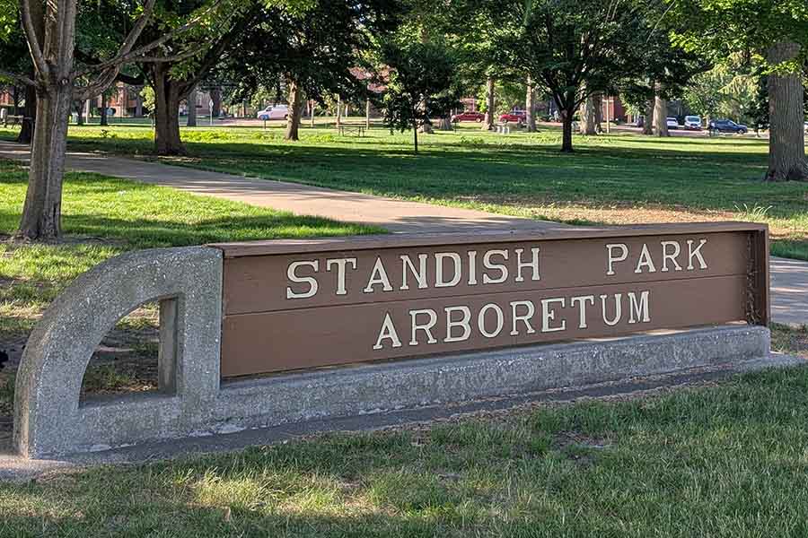 A sign that reads Standish Park Arboretum at the entrance to the outdoor park with trees and a sidewalk in the background.