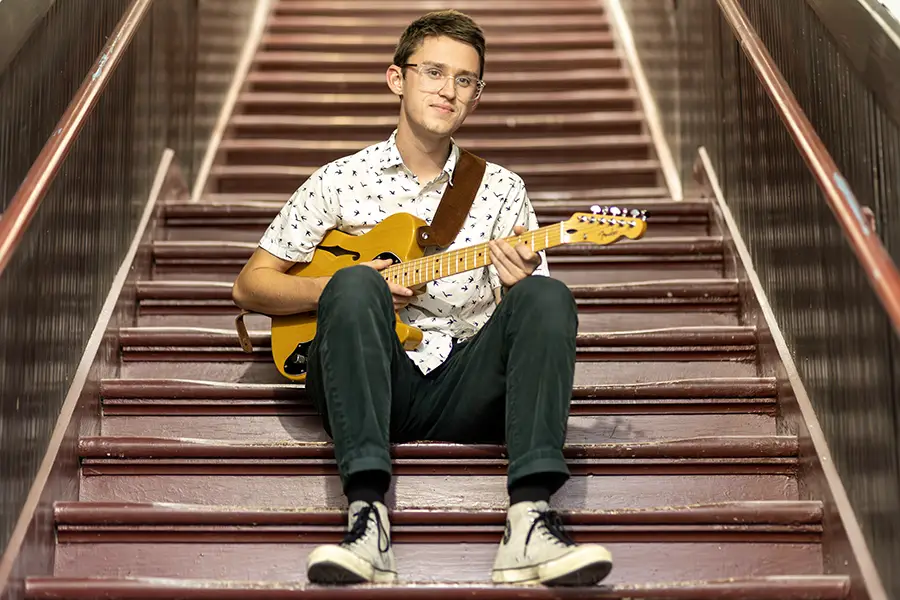 Brandon Roberts ’25 sits on a set of stairs while holding a guitar.