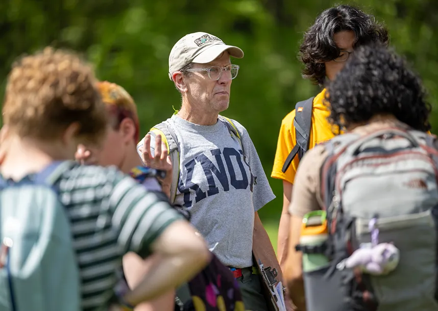 A professor in a Knox t-shirts stands at the center of small group of Knox students, green trees in the background.