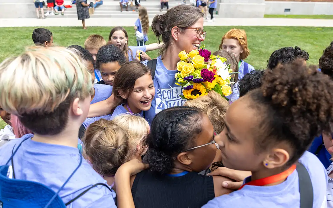 A group of elementary school students surround a  Knox College 4 Kids tteacher, who holds a big bouquet of flowers. 