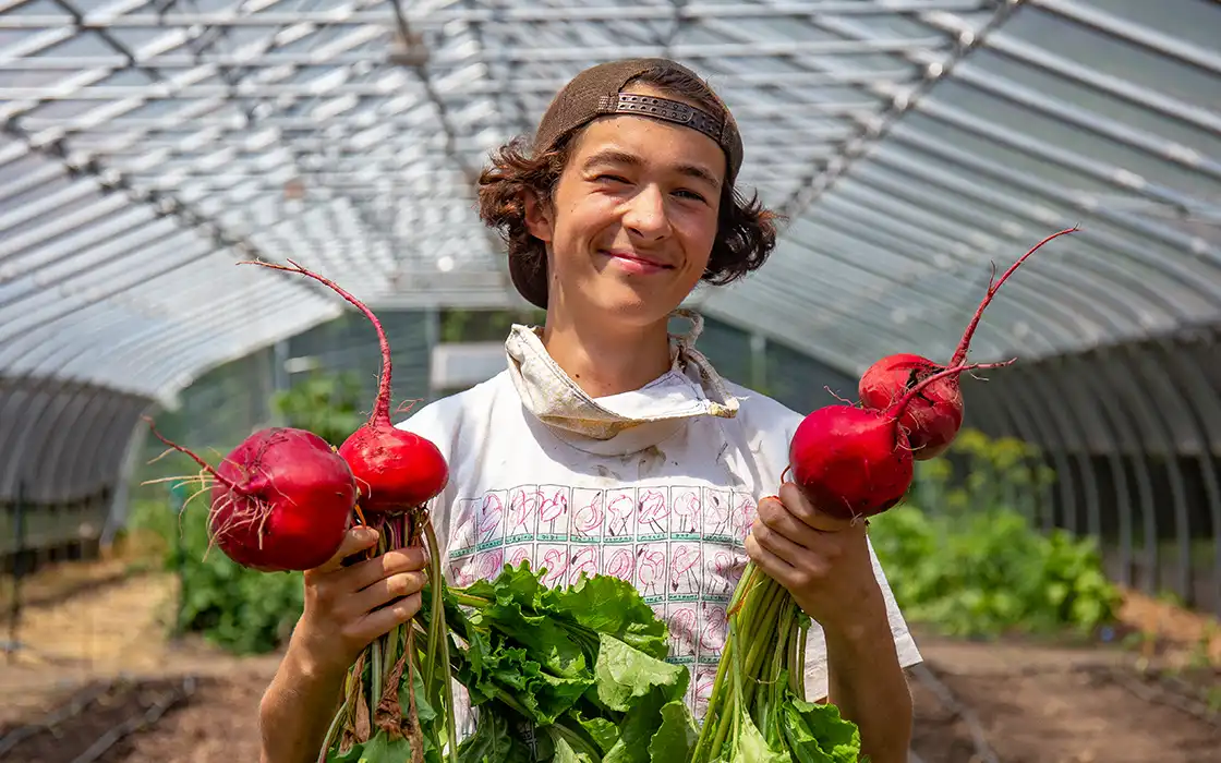 A student smiles at the camera while holding a bunch of red radishes in each hand.