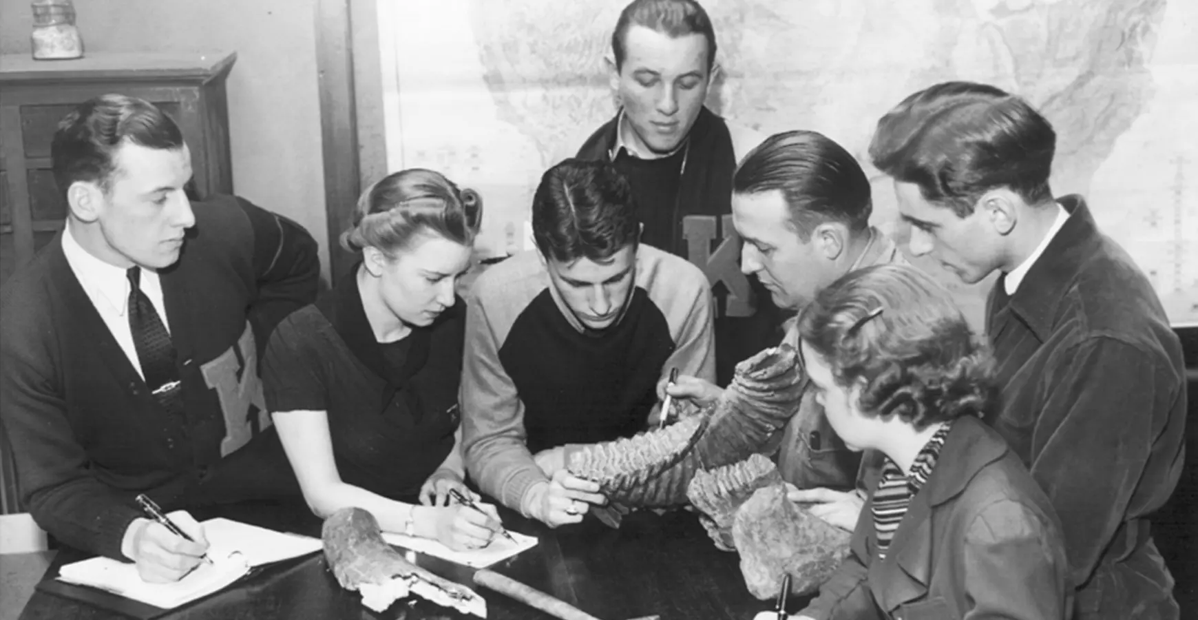A black and white photo from 1957 thar shows a group of students gathered in a circle looking at fossils. 