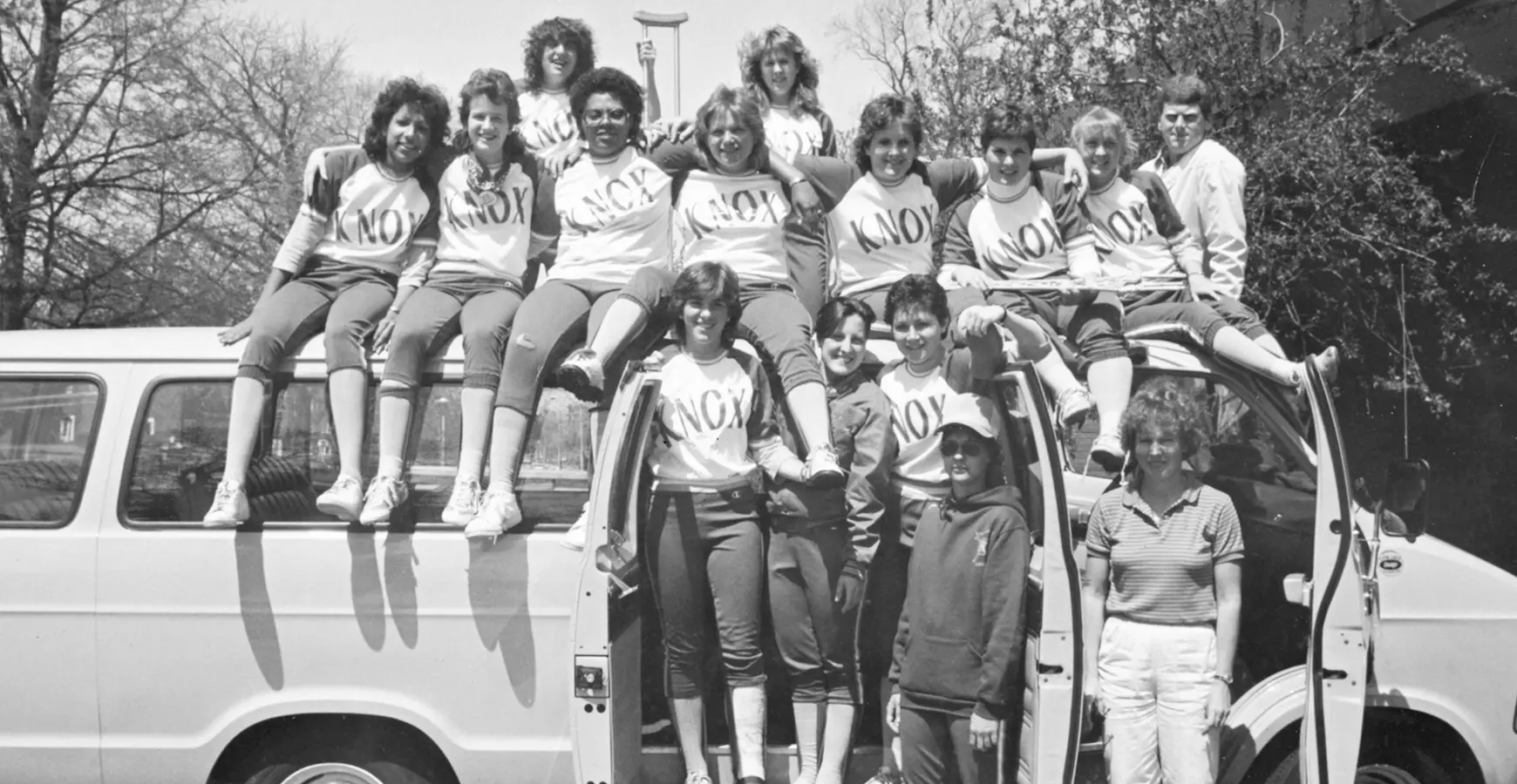 A black and white photo of the Knox College softball team, circa 1980. The team sits on top and stands in front of a white bus