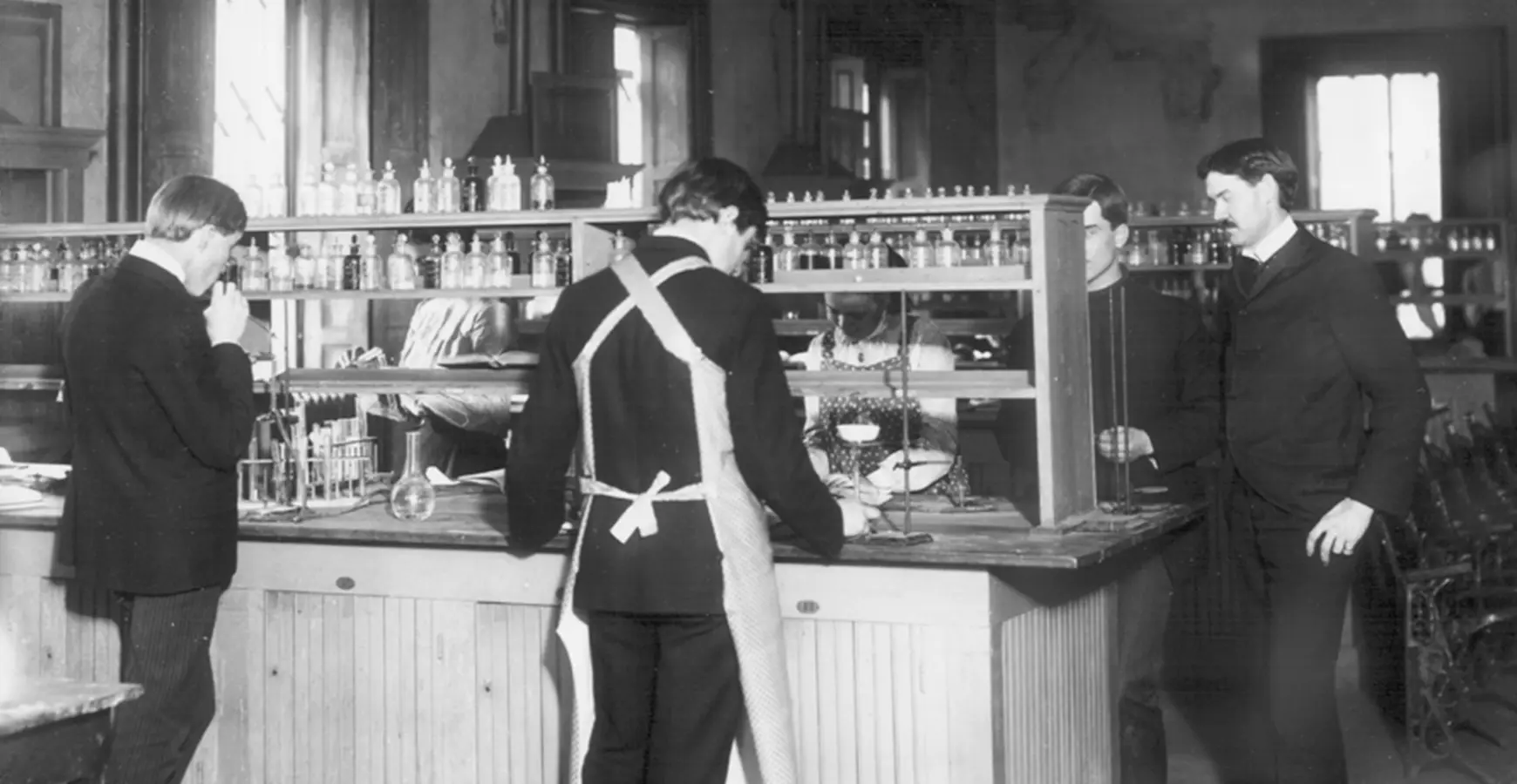 A black and white photo of a chemistry class in the early 20th century. Two students stand at a lab table while a professor oversees their work. 