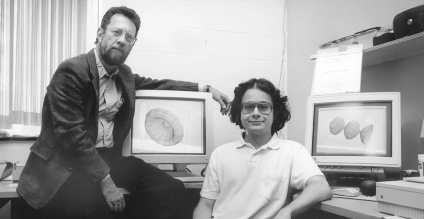 A black and white photo of Professor of Mathematics Dennis Schneider and a student sitting in front of two computer screens featuring graphic models. 