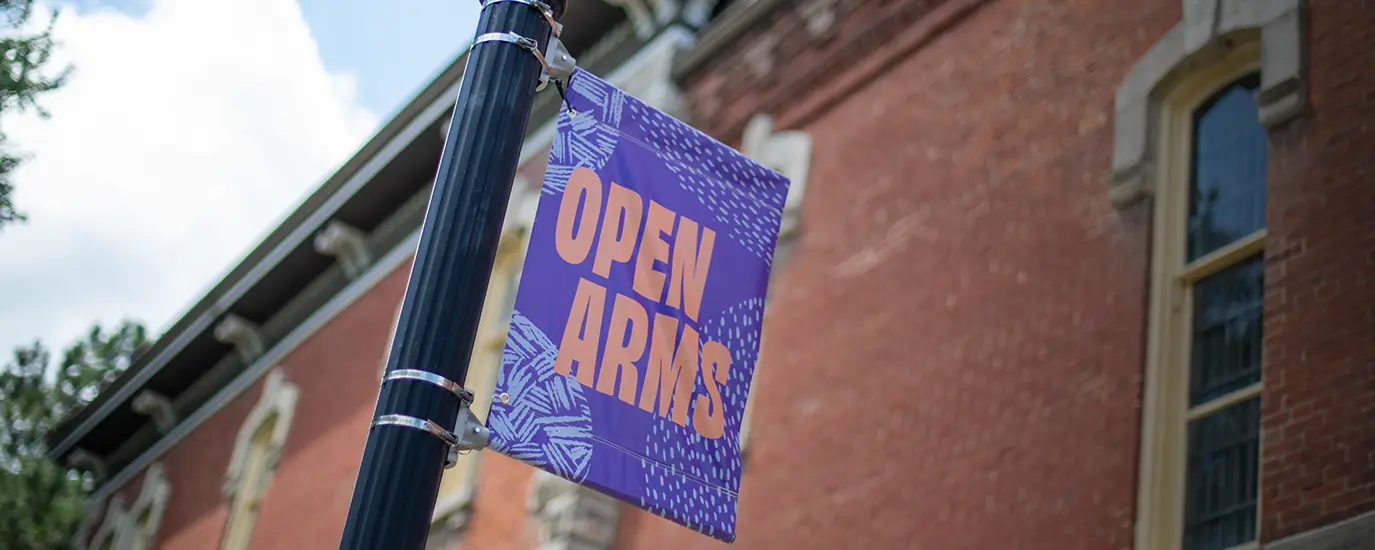 A purple banner that says "Open Arms" on a light pole in front of a brick building.