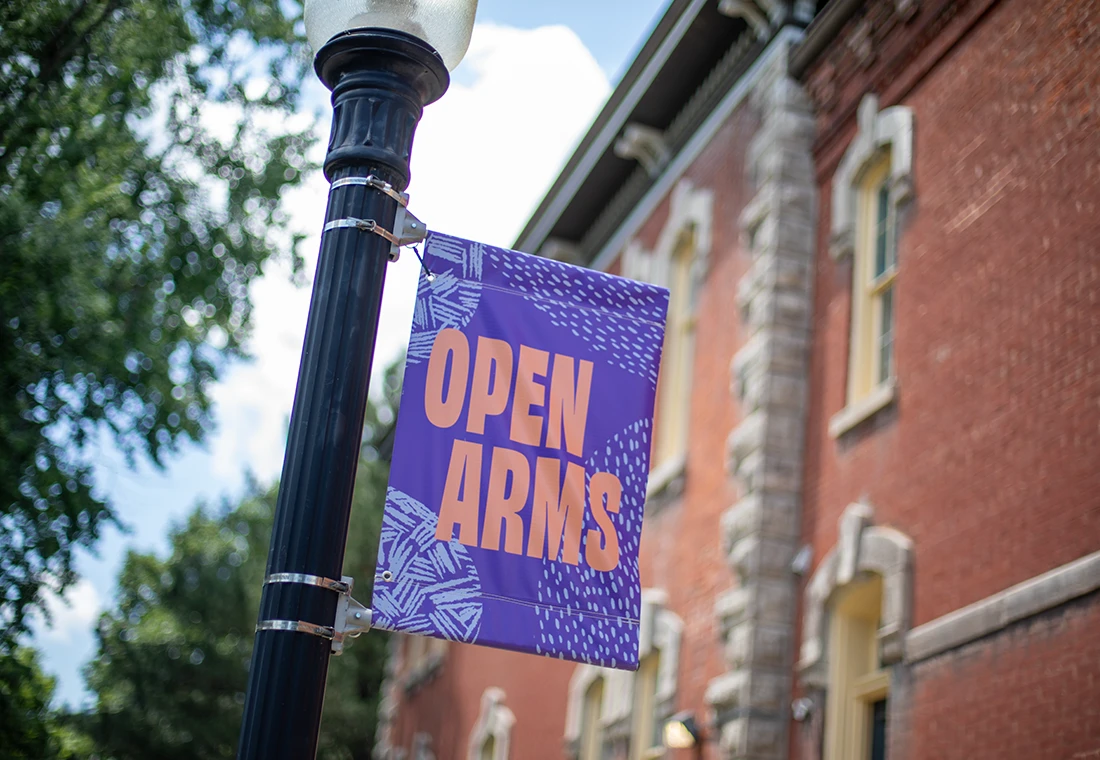 A purple banner on a black light pole. Pink text saying "Open Arms" is featured on the banner.
