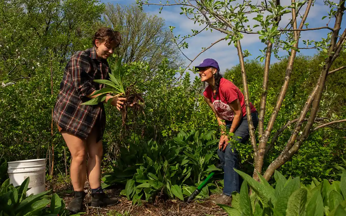 A student holds a plant with its roots exposed while Tina Hope smiles on. They are surrounded by trees and plants.