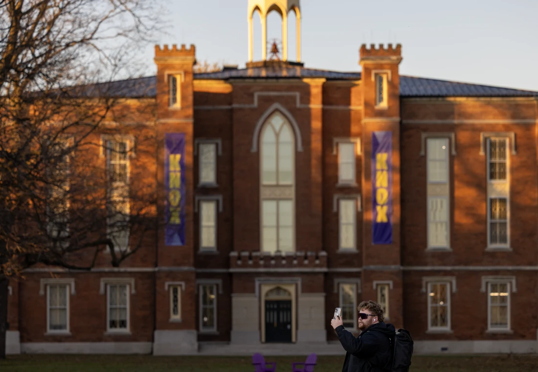 A man takes a selfie on the South Lawn of Knox College's Old Main at sunset.