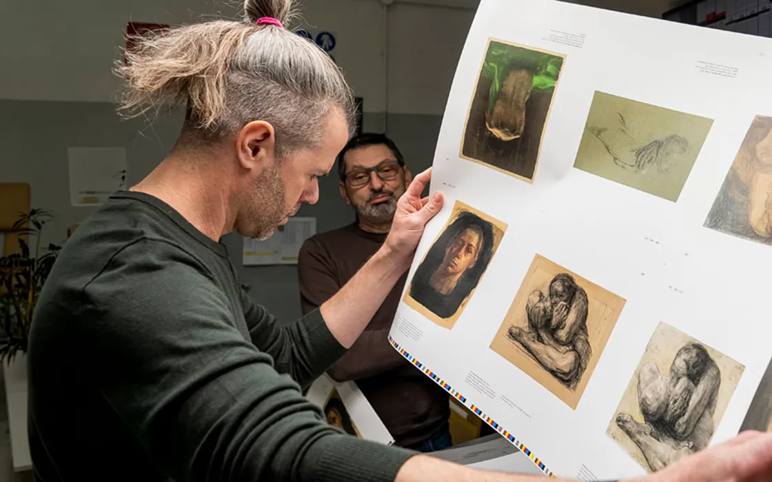 A man scrutinizes images of art printed on a large piece of paper while another man waits in the background.