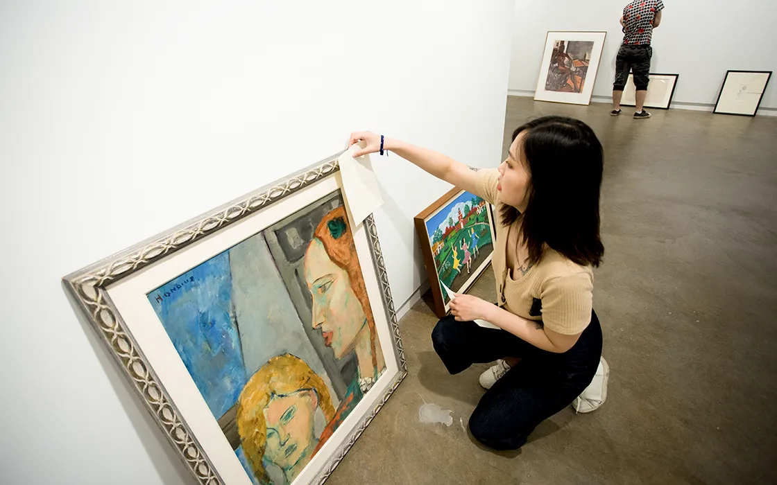In a gallery setting, a student crouches down to affix a piece of paper to a painting.