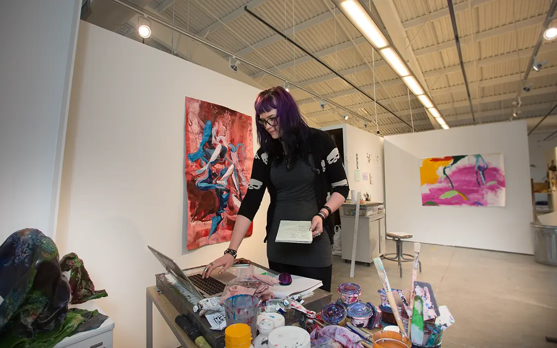 A student works on a laptop in a studio environment. She's surrounded by paintings, paints, and a sculpture.