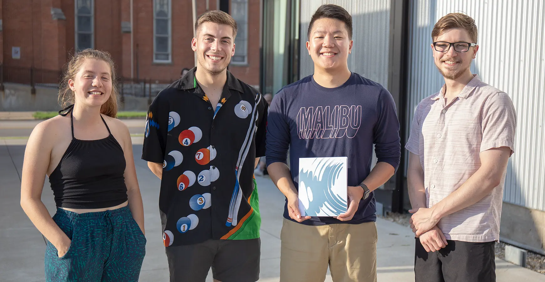 Four students stand outside of the Whitcomb Art Center. One holds a small canvas in his hands with a blue design on it.