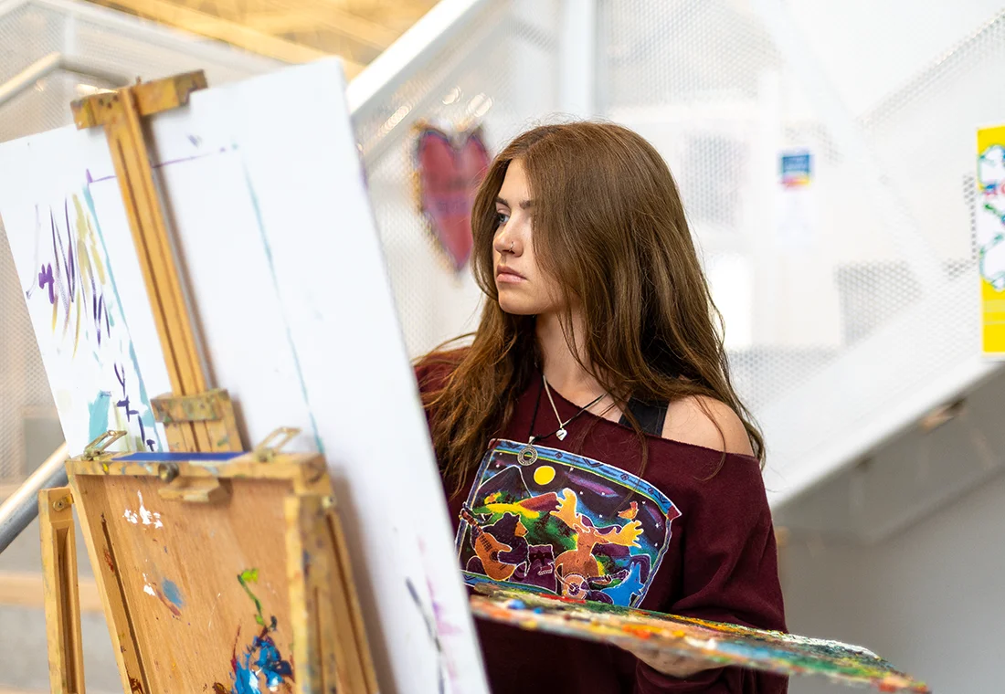Student sits in front of an easel wearing a colorful shirt, painting.