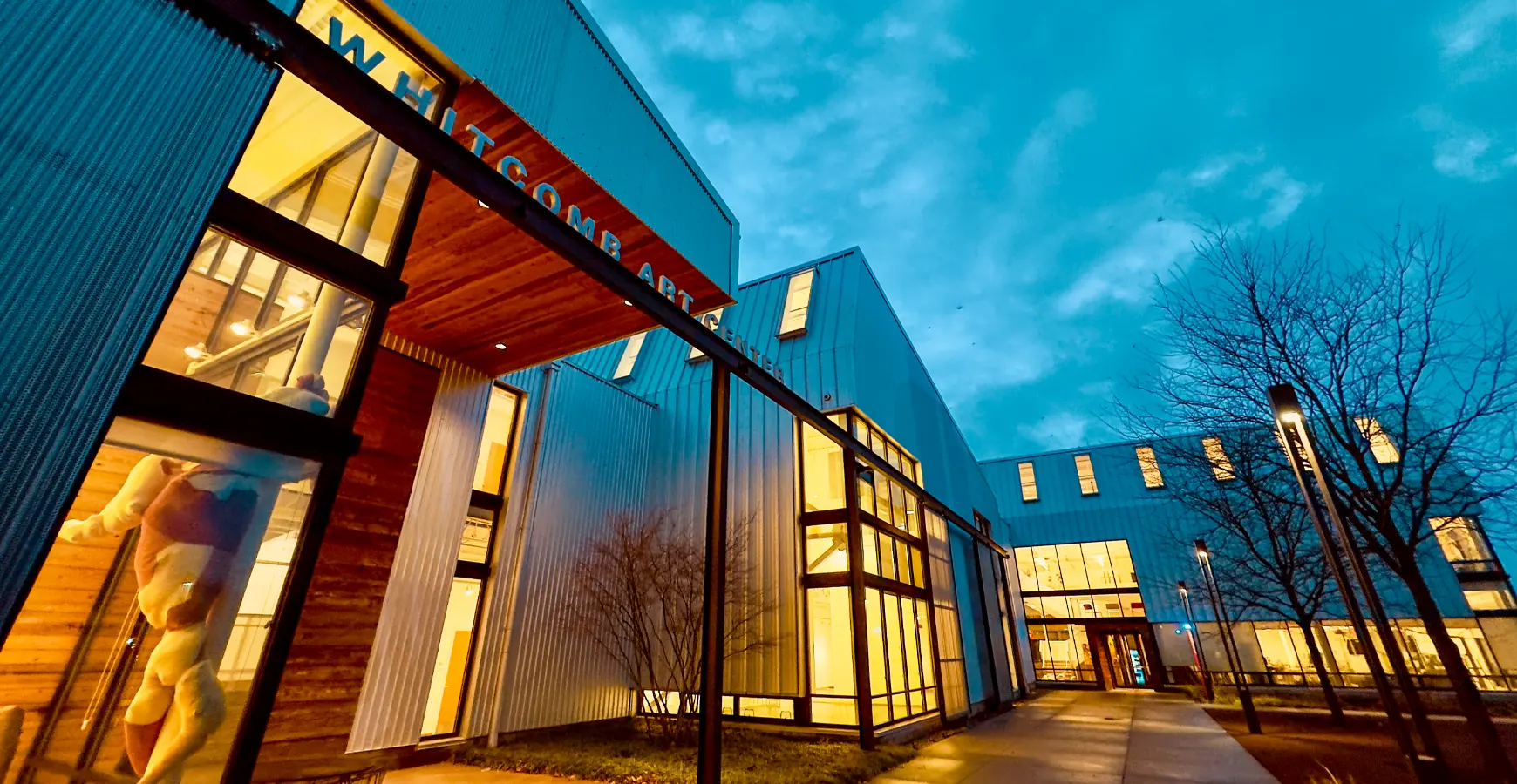 Whitcomb Art Center pictured against a twilight blue cloudy sky, lit with warm yellow lights from within. A sculpture is visible in the nearest window.