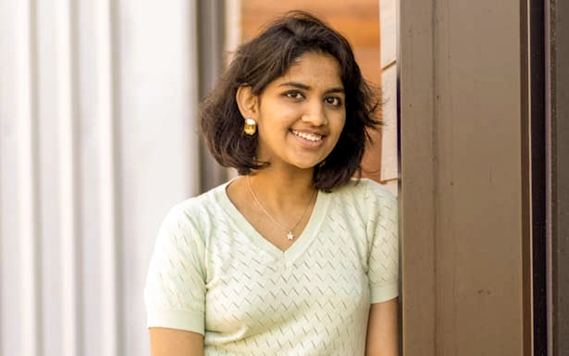 Naysha Jain leans against an exterior wall. Her reflection is faintly visible in the window next to her.
