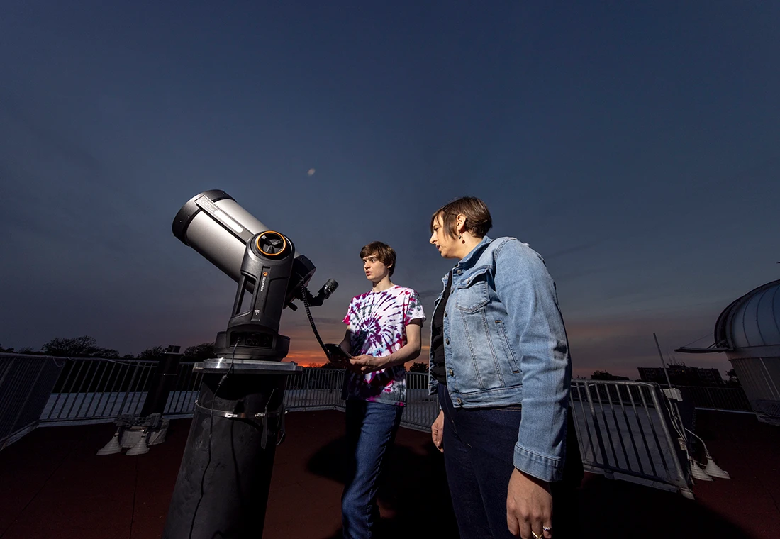 A student and a professor stand on top of the Umbeck Science-Mathematics Center. A telescope is in front of them on a pedestal; a deep blue twilight sky is behind them.