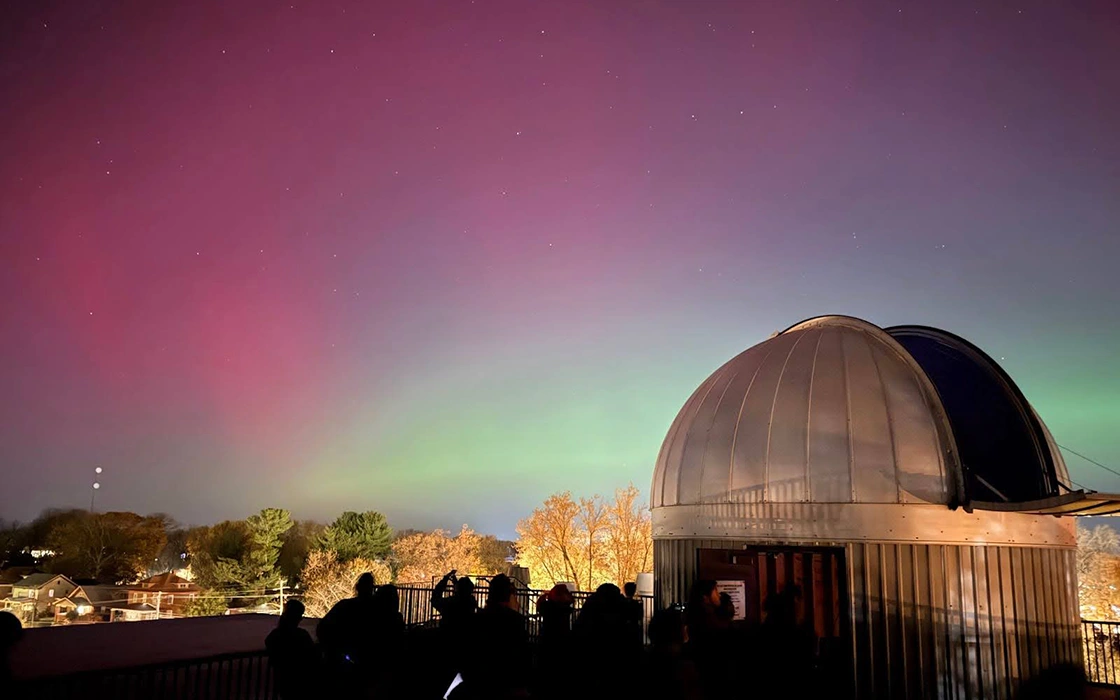 The Knox observatory is on the right. Before it, a colorful sky shows stars and Northern Lights. In the foreground, shadows of students are visible.