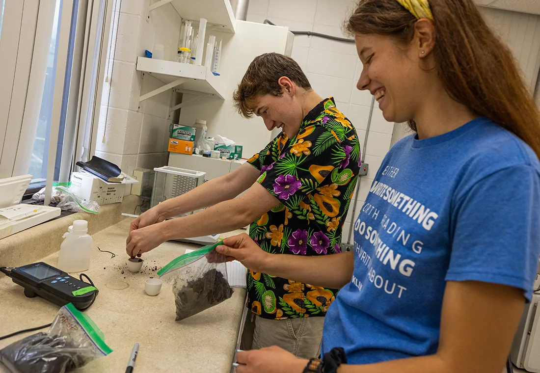 Two students work in a lab. One holds a plastic bag of dirt while the other one places dirt in a small container.
