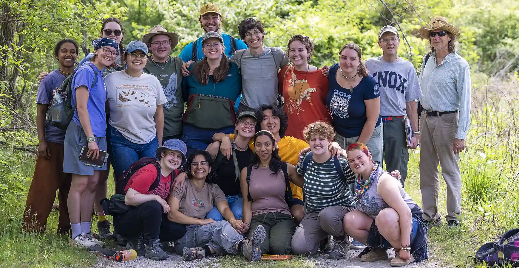 A large group of students and professors smile, surrounded by trees.