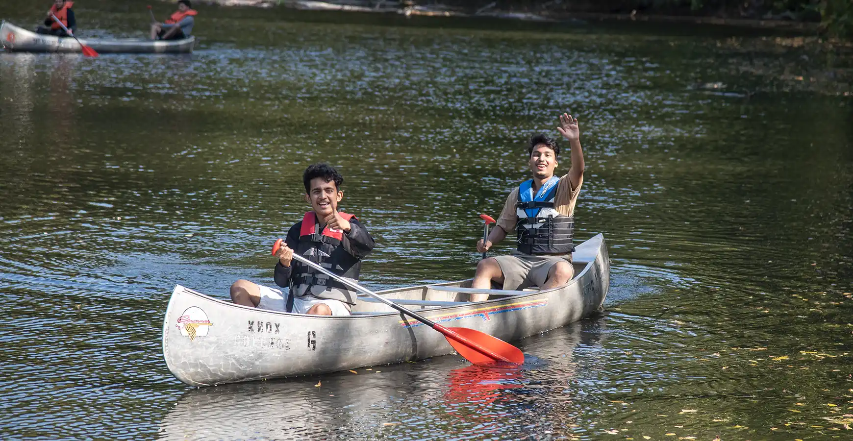 Two students in a kayak smile. One waves, and one gives a thumbs up sign.