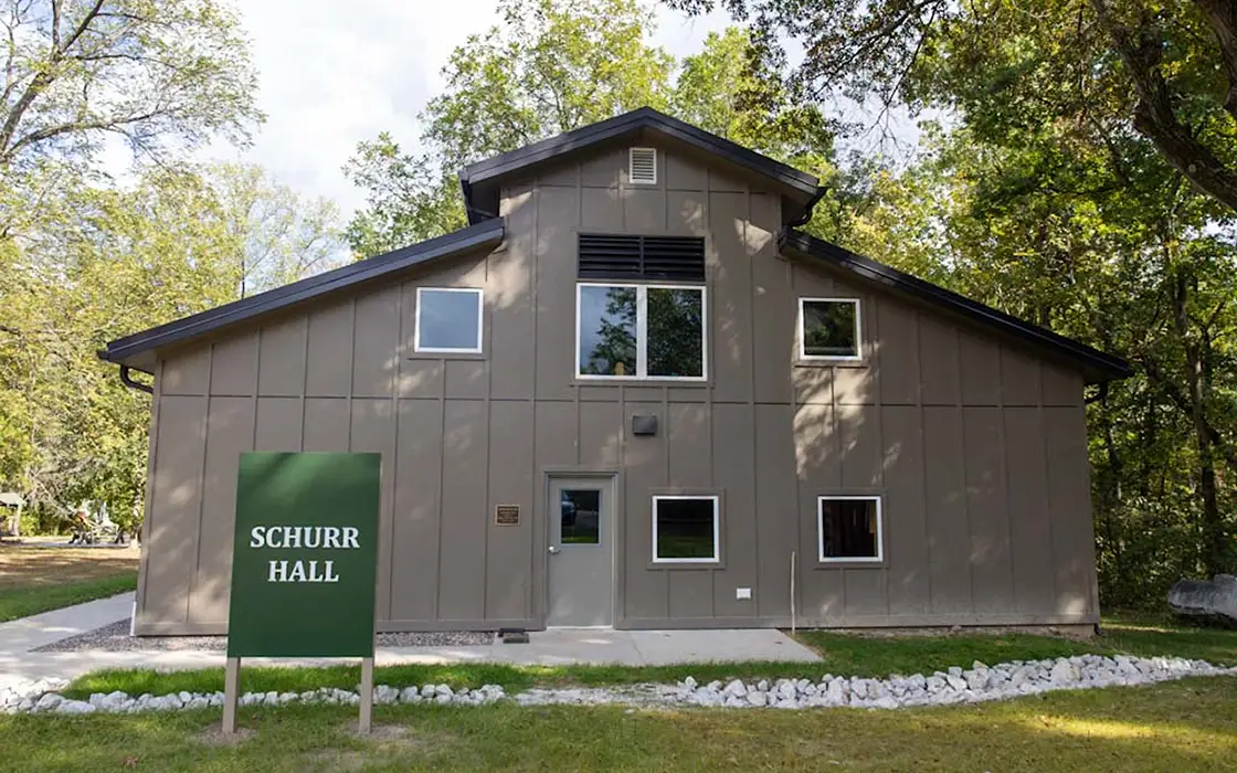 Exterior shot of Schurr Hall, a brown building shaded by trees. The door is visible as well as five windows.